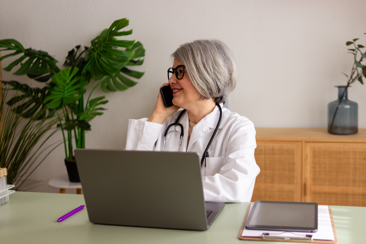 Mature professional doctor talking on smartphone in her office with a laptop and stethoscope