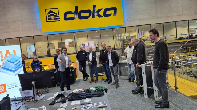 Teilnehmer des Doka Sicherheits-Dialogs stehen in einer Ausstellungshalle rund um eine Präsentationsfläche mit Arbeitskleidung und technischen Exponaten. Im Hintergrund sind Schalungssysteme und ein großes Doka-Logo zu sehen.